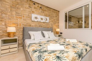 A bedroom with a stone wall, a bed with a floral bedspread, two bedside tables with lamps, and a large window. - Apartment Sealine One (Omis)