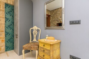 A room with a stone wall, a wooden chair with a leopard print cushion, a wooden dresser with a hat on top, and a mirror on the wall. - Apartment Sealine One (Omis)