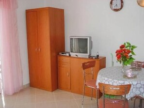 Ein Zimmer mit einem Holzkasten, einem Fernseher darauf, einem Esstisch mit Stühlen und einer Uhr an der Wand.