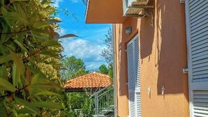 A narrow paved pathway leading to a building with a balcony, a red-tiled roof visible in the background, and a metal gate. - Apartments Eden (Crikvenica)