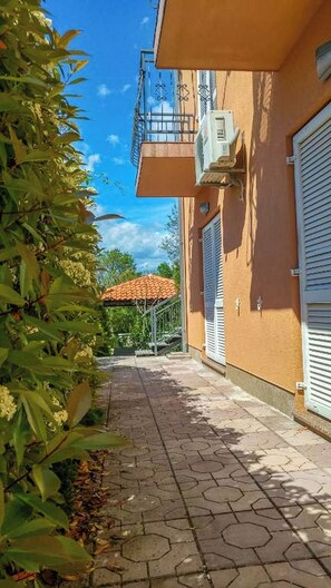 A narrow paved pathway leading to a building with a balcony, a red-tiled roof visible in the background, and a metal gate. - Apartments Eden (Crikvenica)