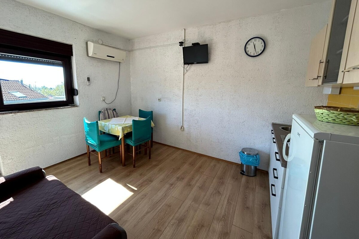 A compact room with a dining table, chairs, a TV, a clock, a washing machine, and a window.