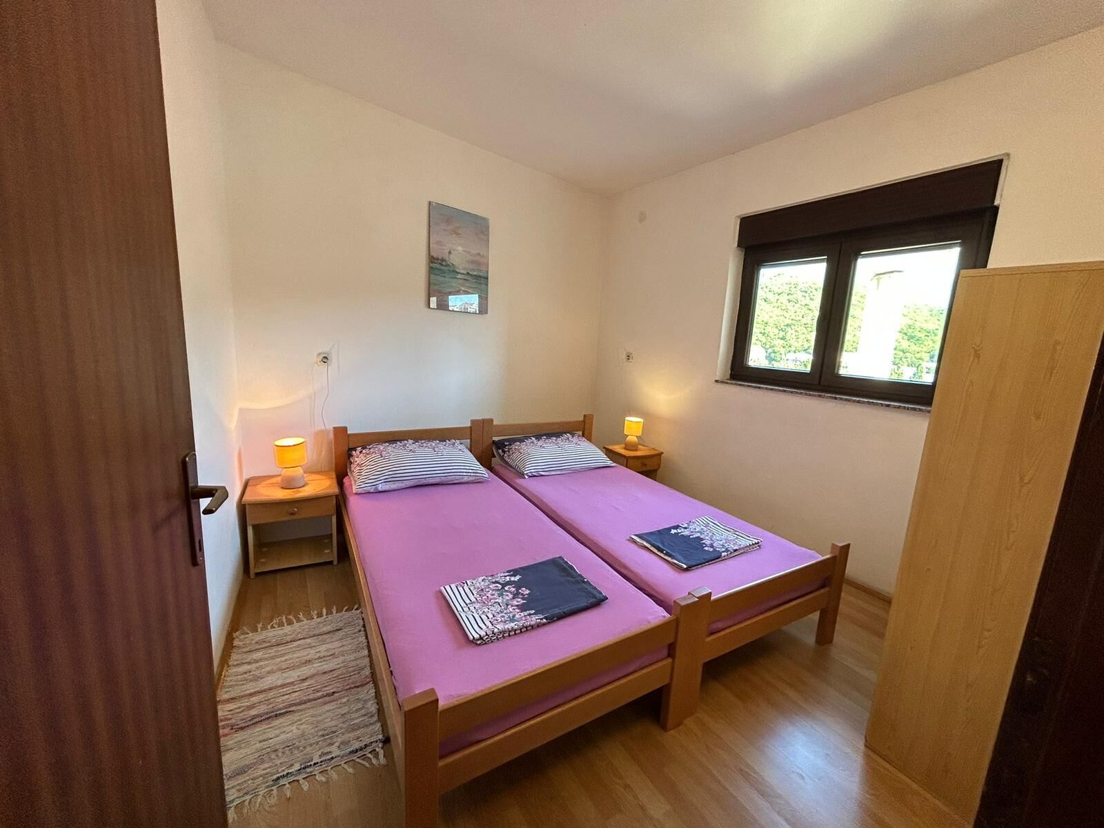 A small, well-lit room with two wooden beds, a window with a view, and a painting on the wall.