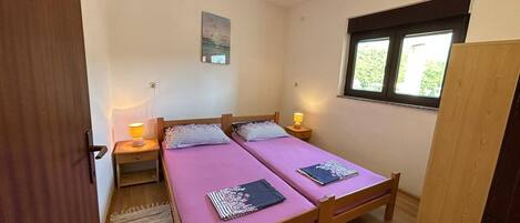 A small, well-lit room with two wooden beds, a window with a view, and a painting on the wall.