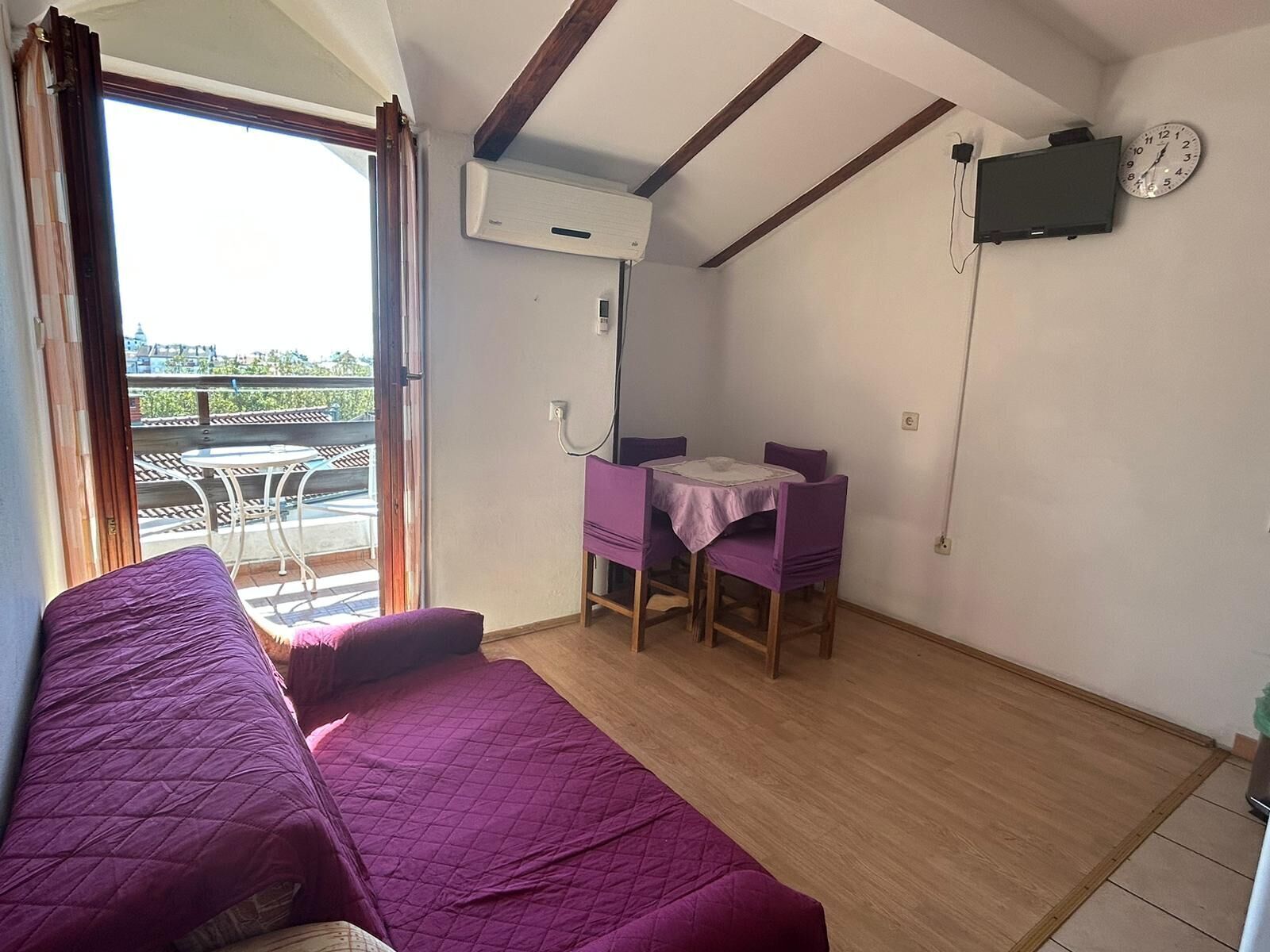 Ein Zimmer mit Bett, einem kleinen Tisch mit Stühlen, einer Klimaanlage, einer Uhr und Blick ins Freie.