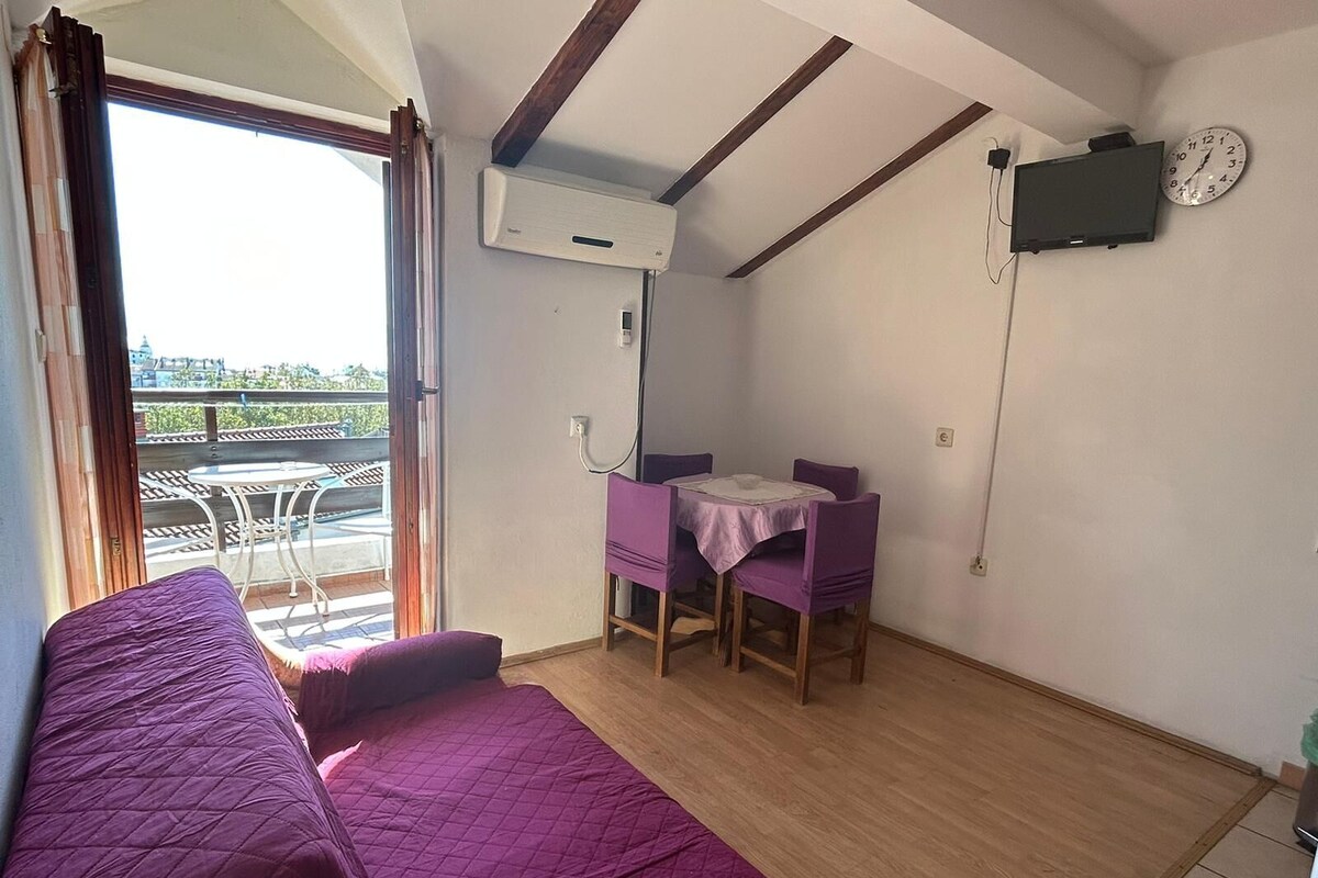 A room with a bed, a small table with chairs, an air conditioning unit, a clock, and a view of the outside.