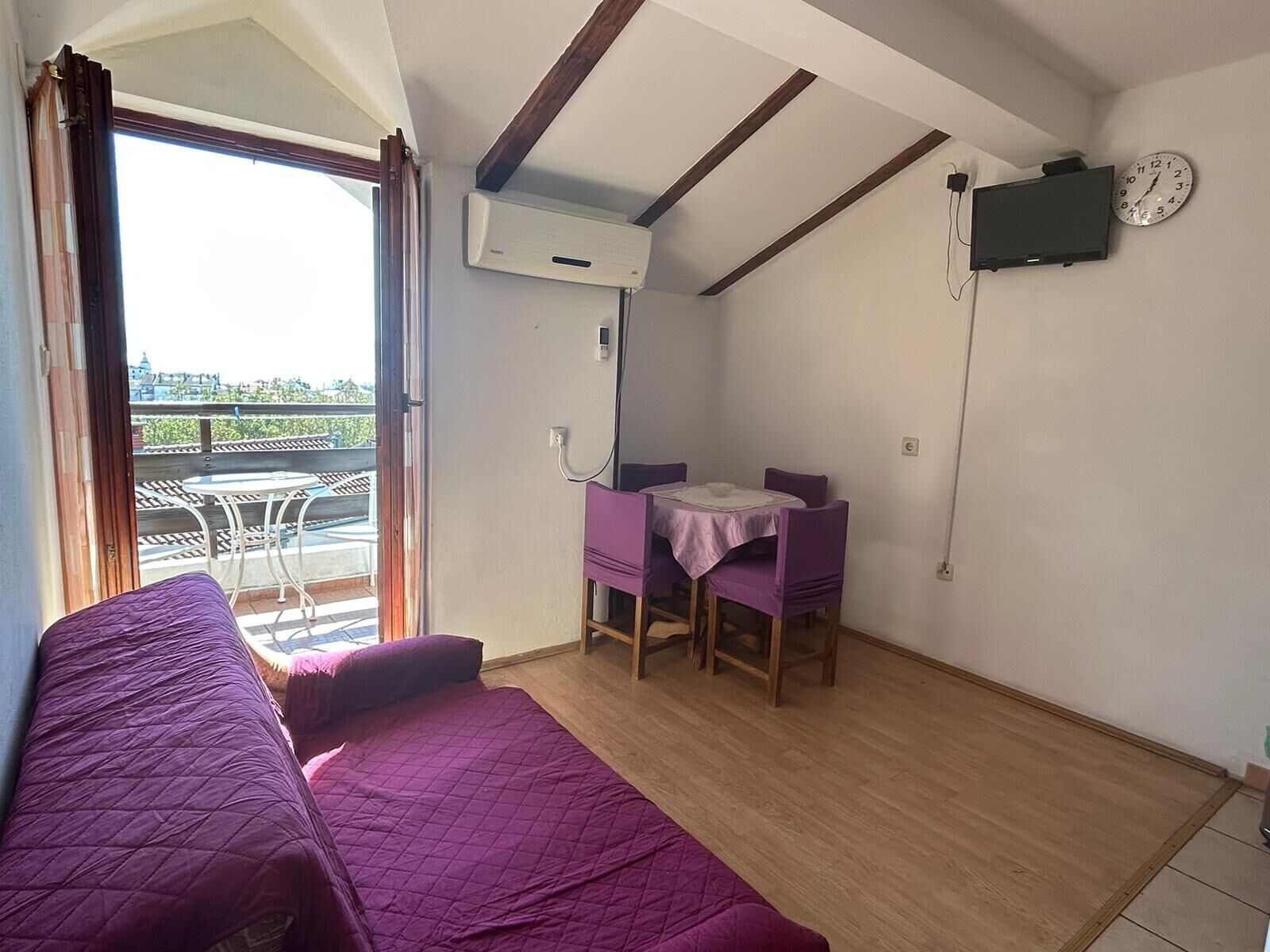 A room with a bed, a table with chairs, an air conditioning unit, a clock, and a view of the outside.