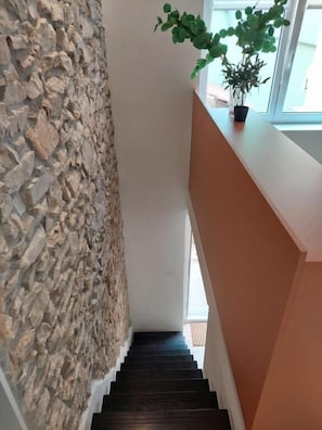 Un escalier avec un mur en pierre d’un côté et une rampe en bois de l’autre, menant à un espace intérieur lumineux.