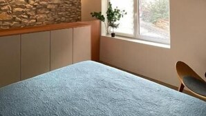 A bedroom with a stone wall, a window with a plant, a bed, and a chair.