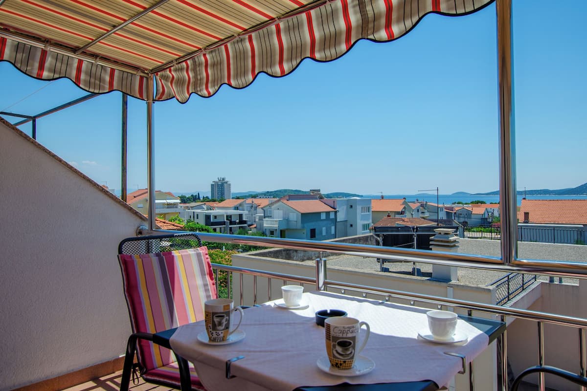 A balcony with a table set for two, a striped chair, and a view of buildings and hills.