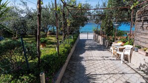 Ein gepflasterter Weg führt zu einer Holzpergola mit Tisch und Stühlen, umgeben von Grün und Blick aufs Meer.
