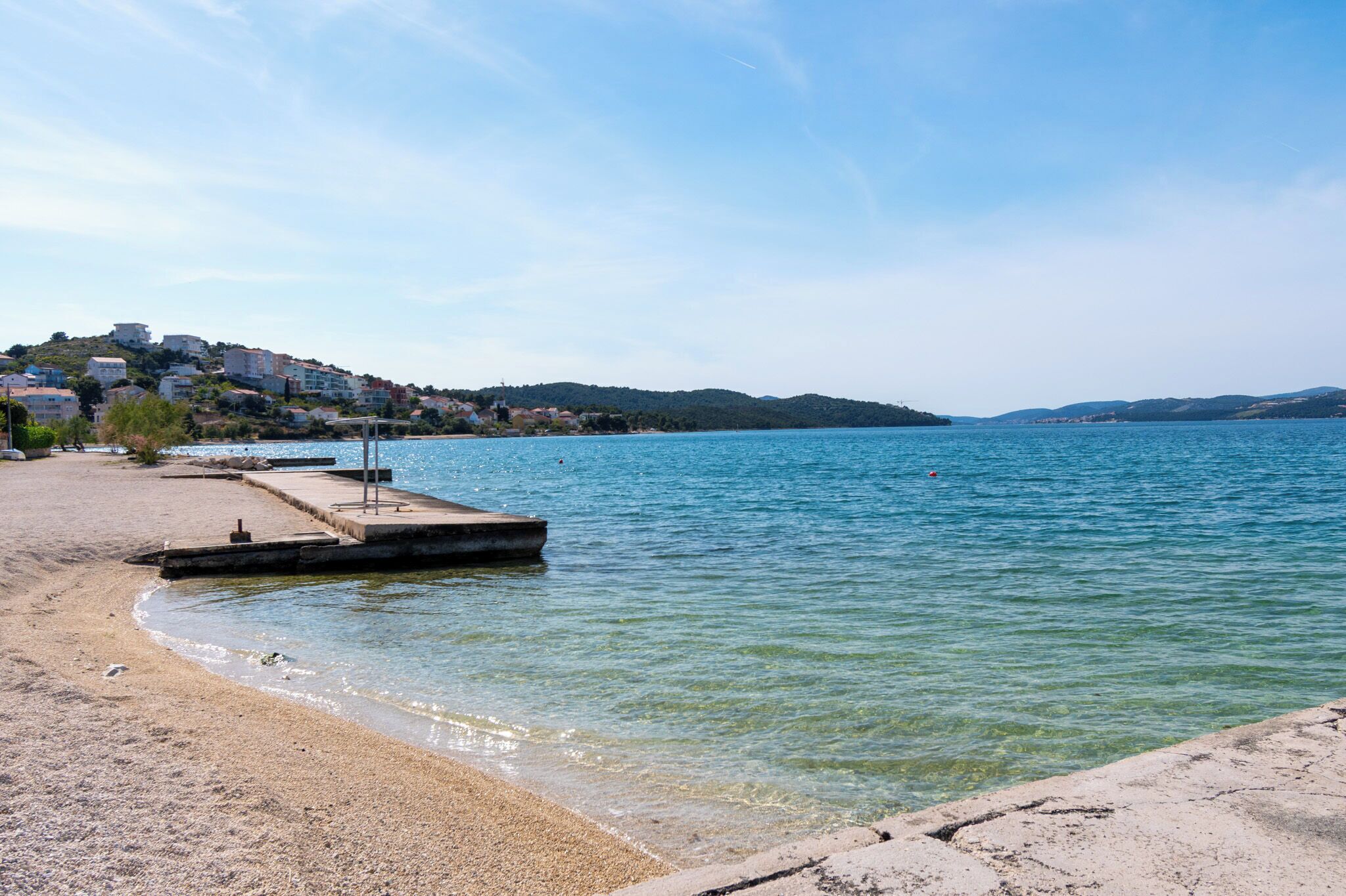 Une plage avec une jetée en béton qui s’avance dans une étendue d’eau calme, entourée de bâtiments et de collines en arrière-plan.