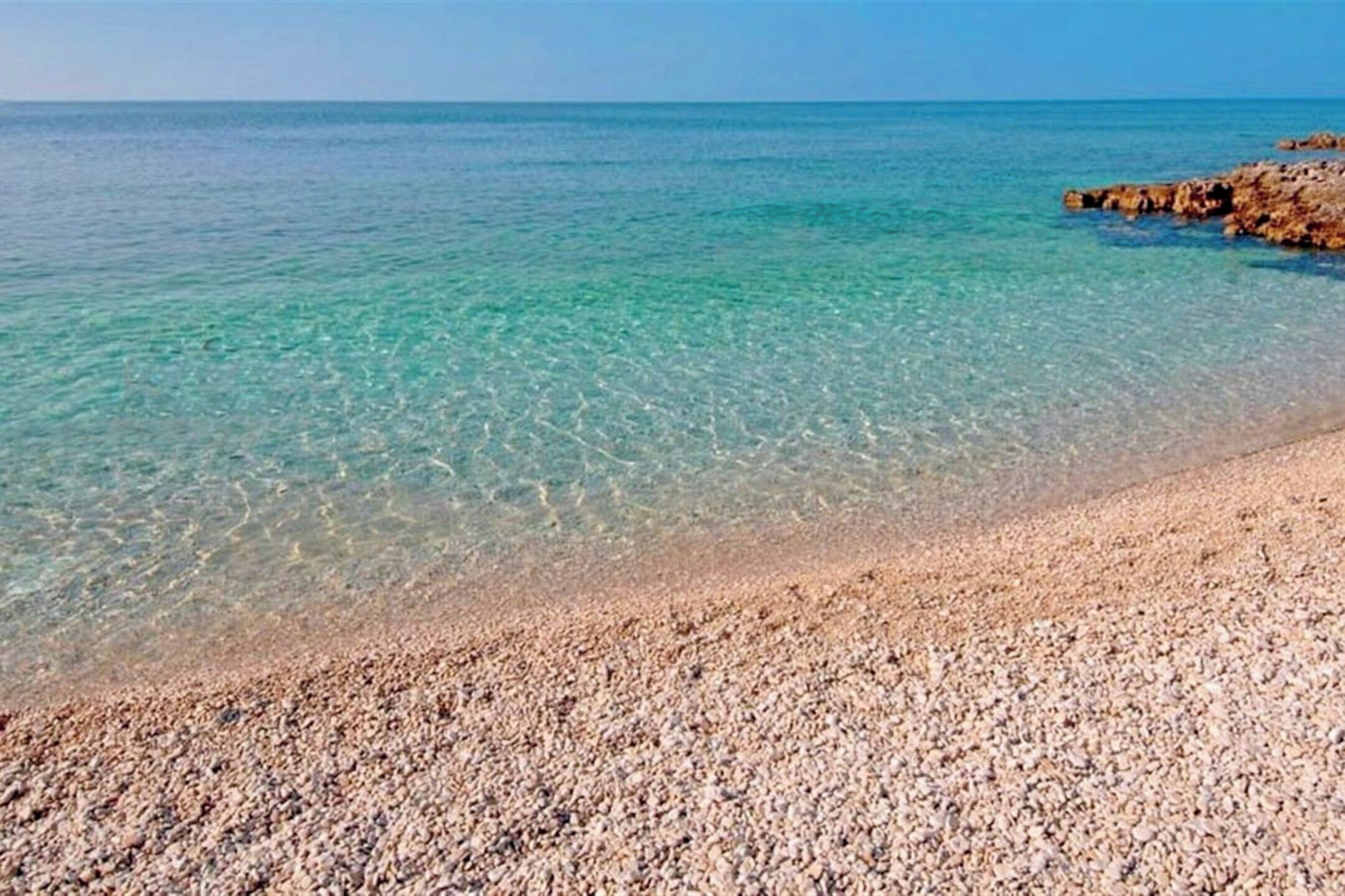 Una playa de guijarros con agua turquesa cristalina y un promontorio rocoso.