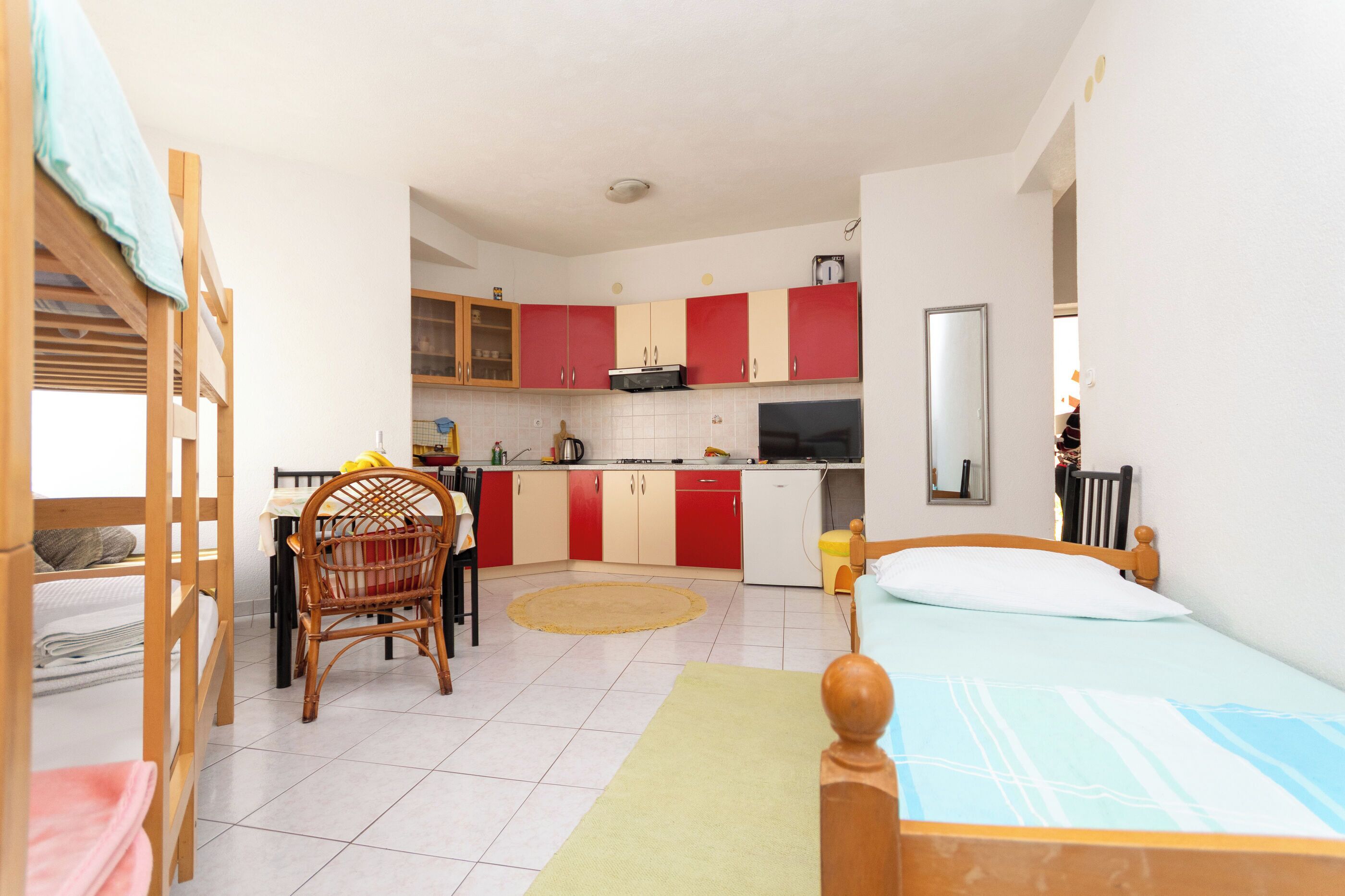 Una habitación funcional con literas, una mesa de comedor con sillas, una cocina con armarios rojos y una pequeña alfombra.
