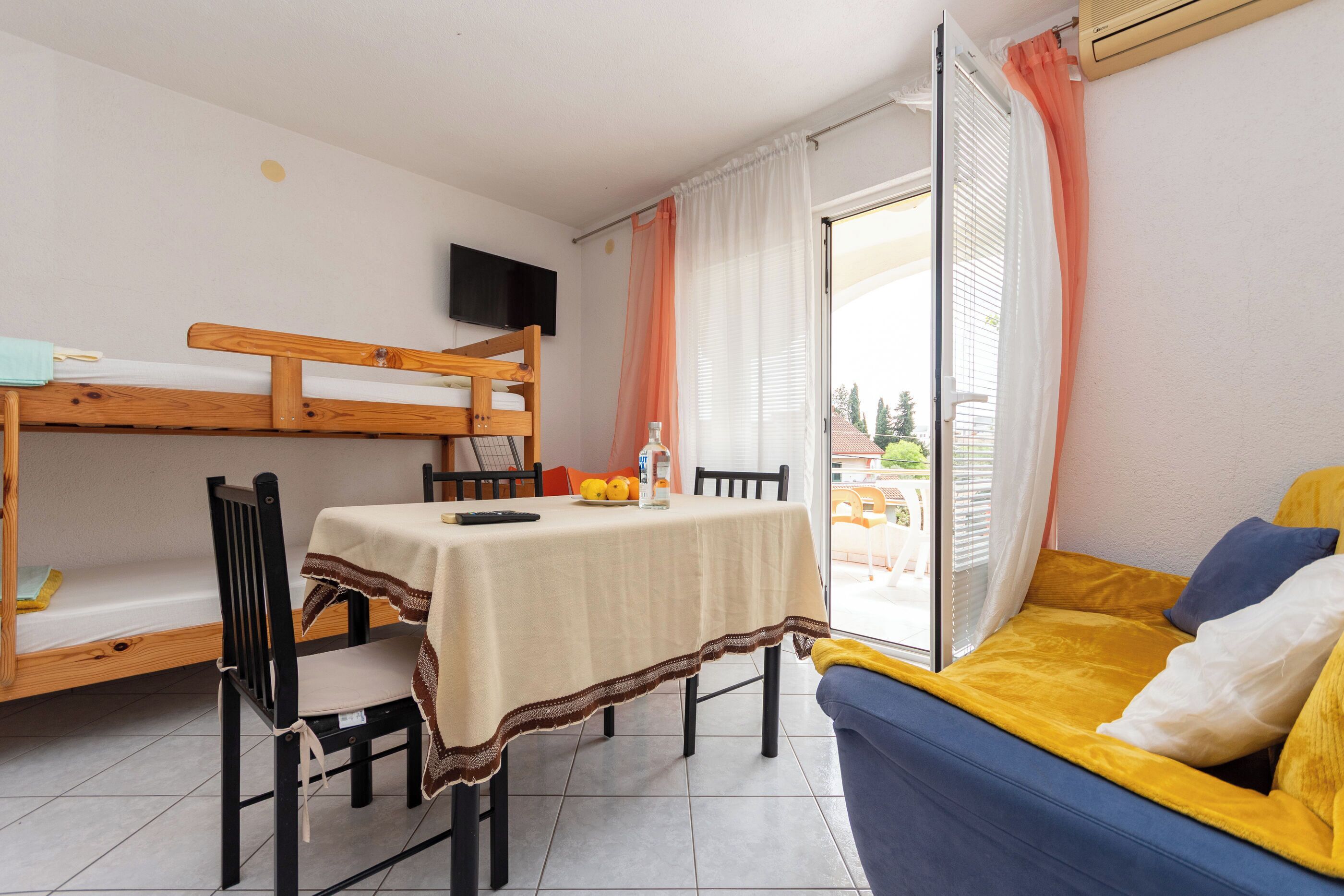 A room with a bunk bed, a table with a tablecloth, a sofa, and a TV mounted on the wall.