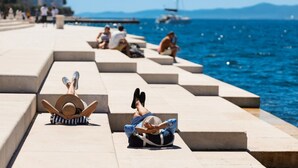 A seaside promenade with steps leading to the water, a person reclining on the steps, and a sailboat in the distance.