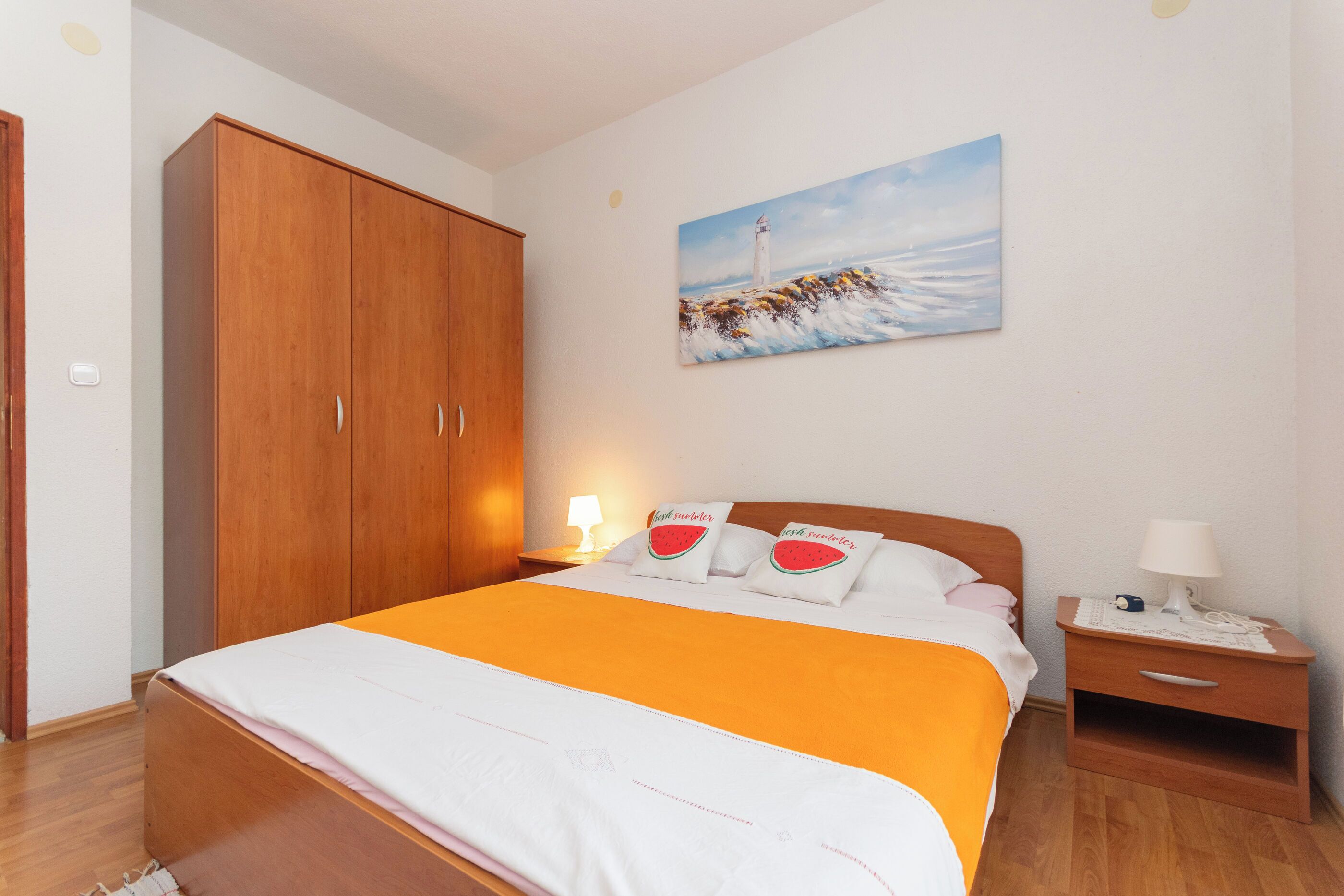 A bedroom with a wooden wardrobe, a bed with an orange bedspread, a nightstand with a lamp, and a painting of a lighthouse on the wall.