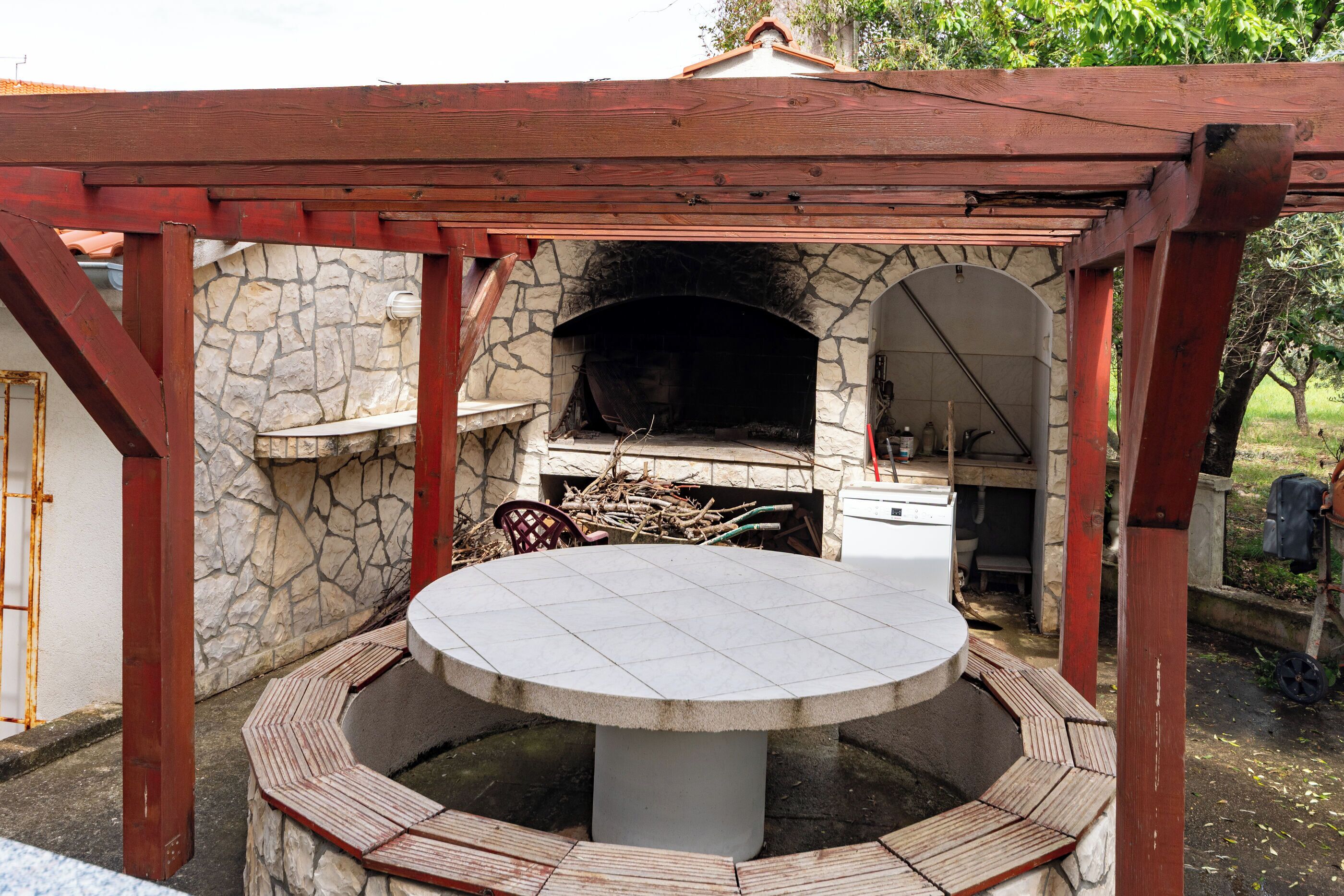Cocina al aire libre de piedra con horno de pizza, una mesa redonda y una estructura de pérgola de madera.