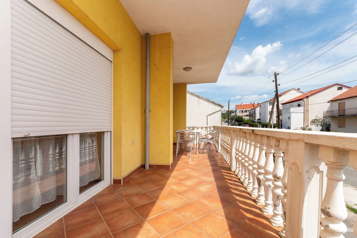 Un balcón con barandilla blanca, piso de baldosas y vista a casas vecinas.