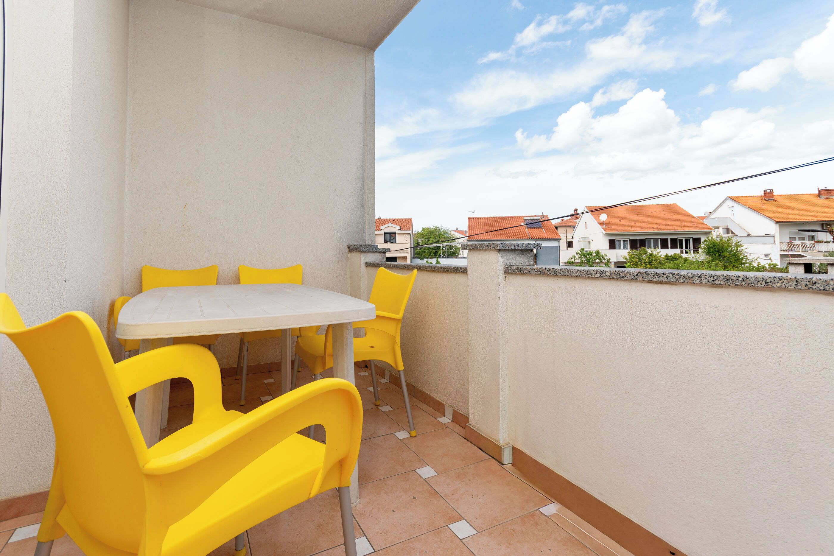 Varanda com uma mesa e cadeiras amarelas, parede branca e vista de casas ao fundo.