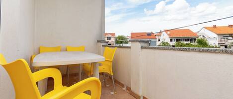 Un balcón con una mesa y sillas amarillas, una pared blanca y vistas a casas al fondo.