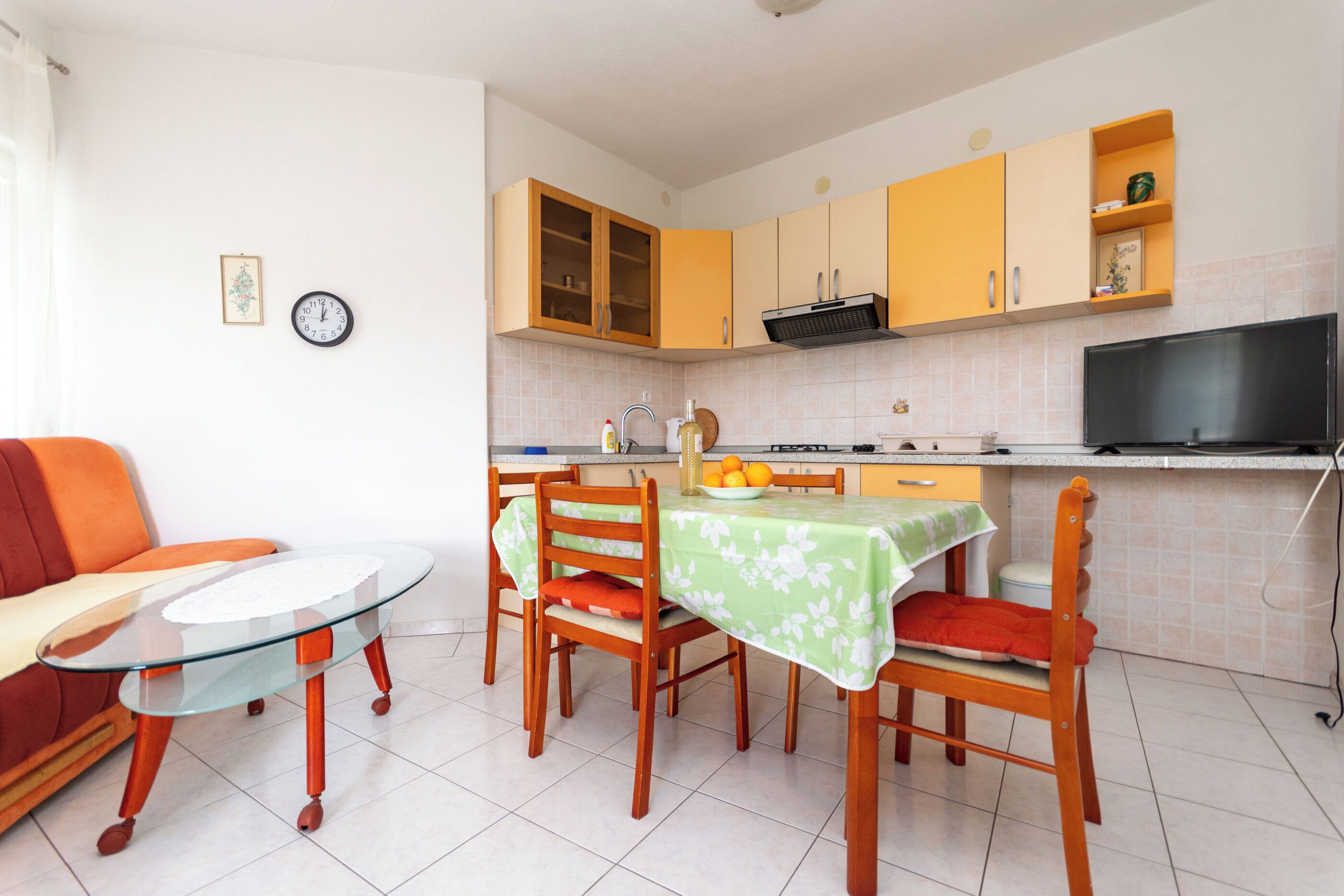 A compact kitchen with wooden cabinets, a dining table with chairs, a small TV, and a clock on the wall.