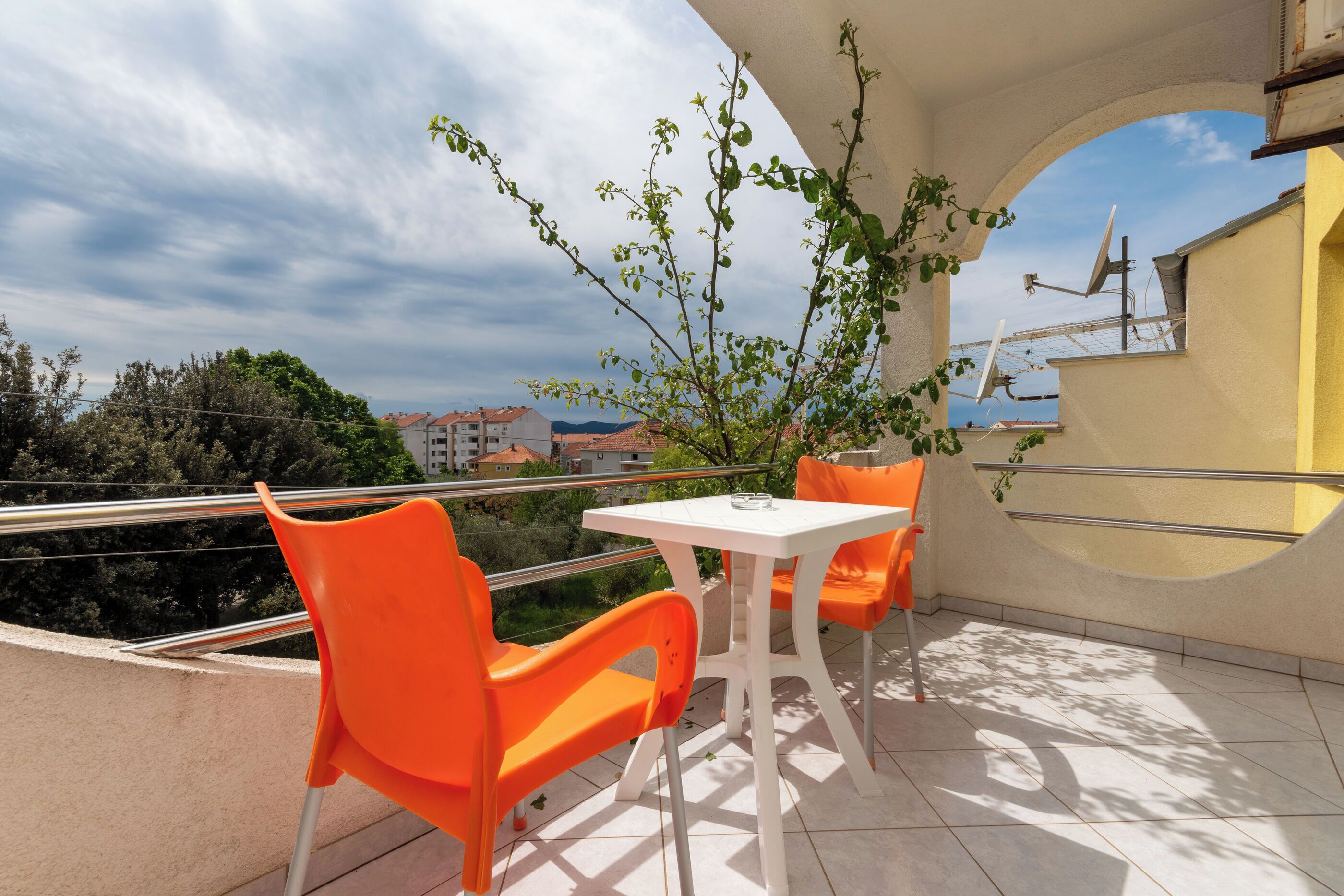 A balcony with two orange chairs and a white table, a potted plant, and a view of buildings and trees.