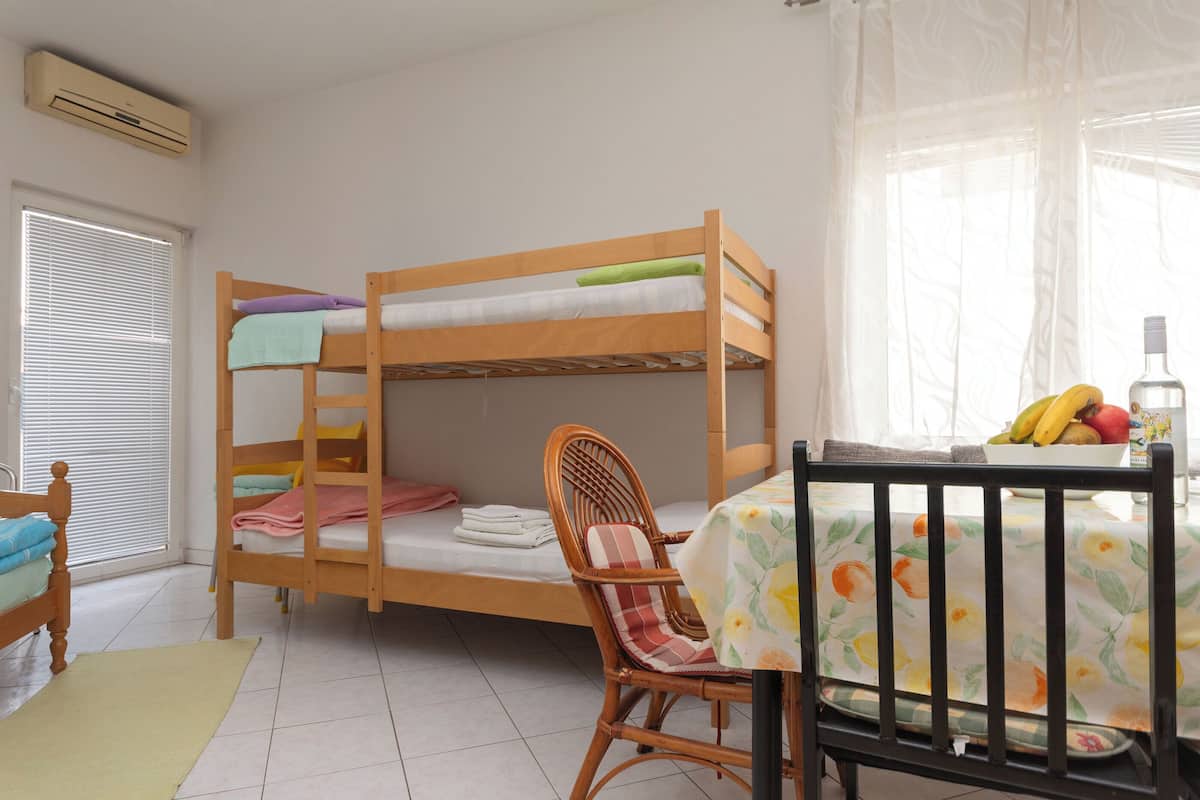 Una cama individual con almohadas, una silla de mimbre, una mesa de comedor con un frutero y una ventana con persianas.