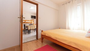 A bedroom with a wooden bed, a red rug, a dining area with a table and chairs, and a television in the background. - Apartments Gardelin in Zadar (Zadar)