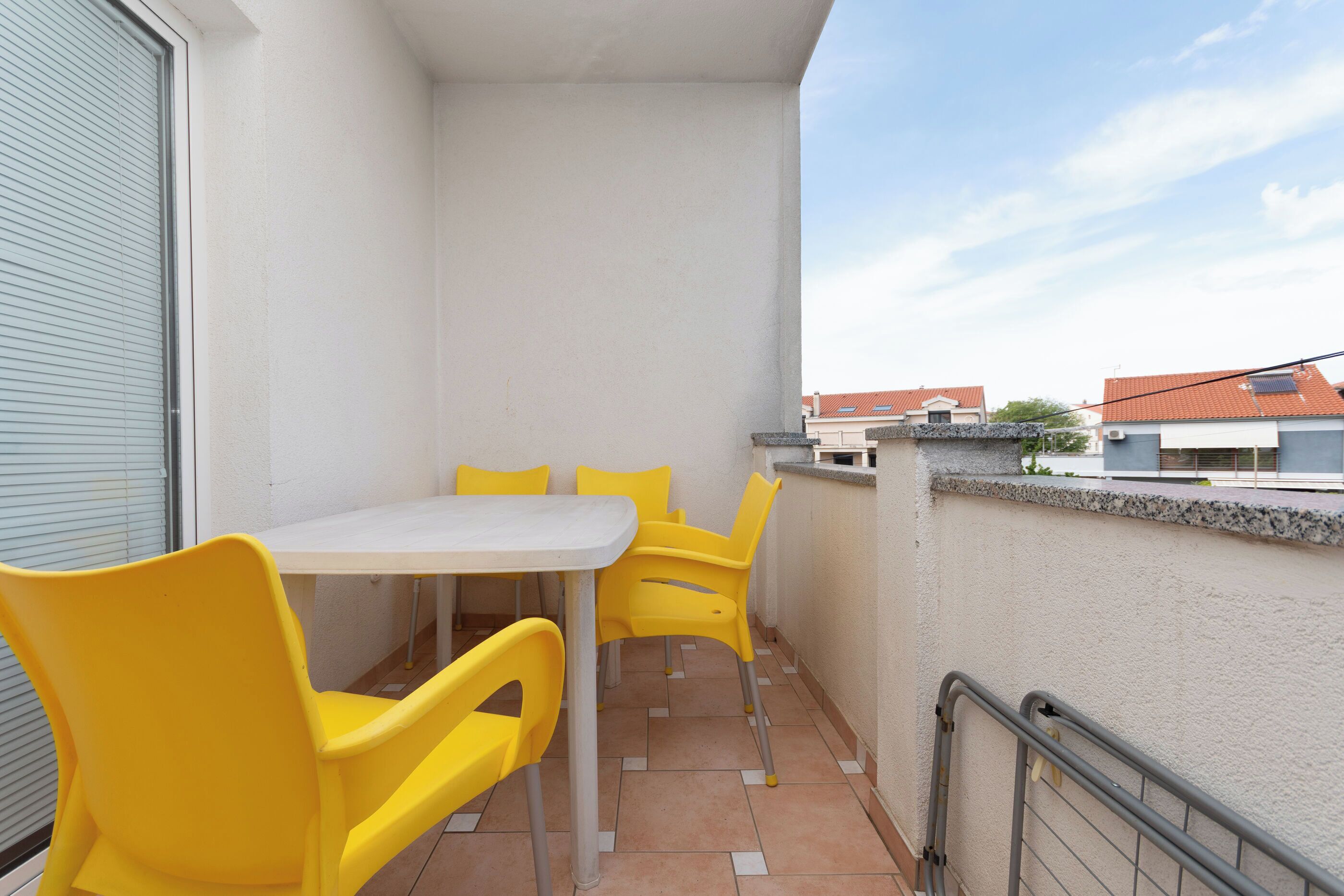 A balcony with a white table and four yellow chairs, a metal railing, and a view of buildings in the background.