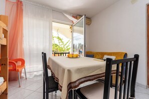 A room with a bed, a table with a tablecloth, a chair, a window with curtains, and a view of buildings outside.