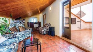 Zona d'estar àmplia amb sostre de fusta, un sofà, una cadira, taula de vidre i una porta corredissa de vidre que dona a zona exterior.