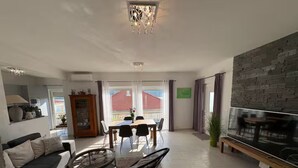 A modern living room with a sofa, coffee table, dining table, and chairs. There is a TV on the wall and a chandelier.