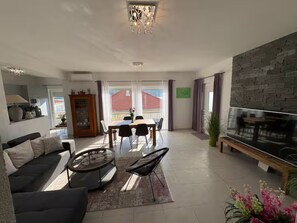 Ein modernes Wohnzimmer mit einer Couch, Couchtisch, Esstisch und Stühlen. An der Wand befindet sich ein Fernseher und ein Kronleuchter.