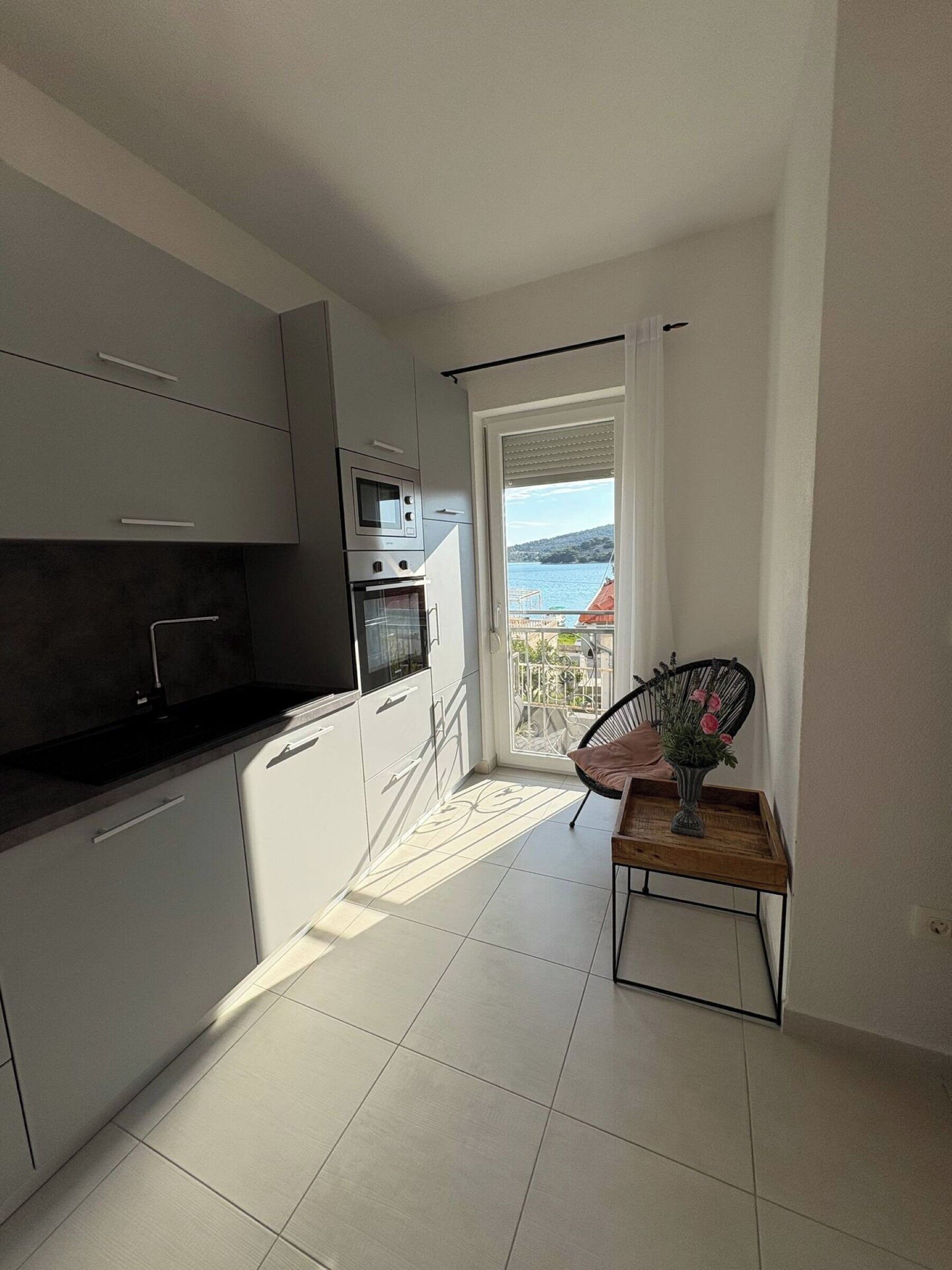 A modern kitchen with a built-in oven and microwave, a dining area with a table and chair, and a balcony with a view of the sea.