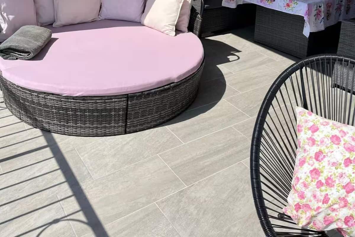Un área de estar al aire libre iluminada por el sol, con un sofá de mimbre redondo, una mesa con mantel floral y una silla de mimbre.