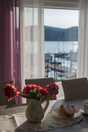 A table with a vase of flowers, a plate with food, and a cup, with a view of a marina and mountains in the background.