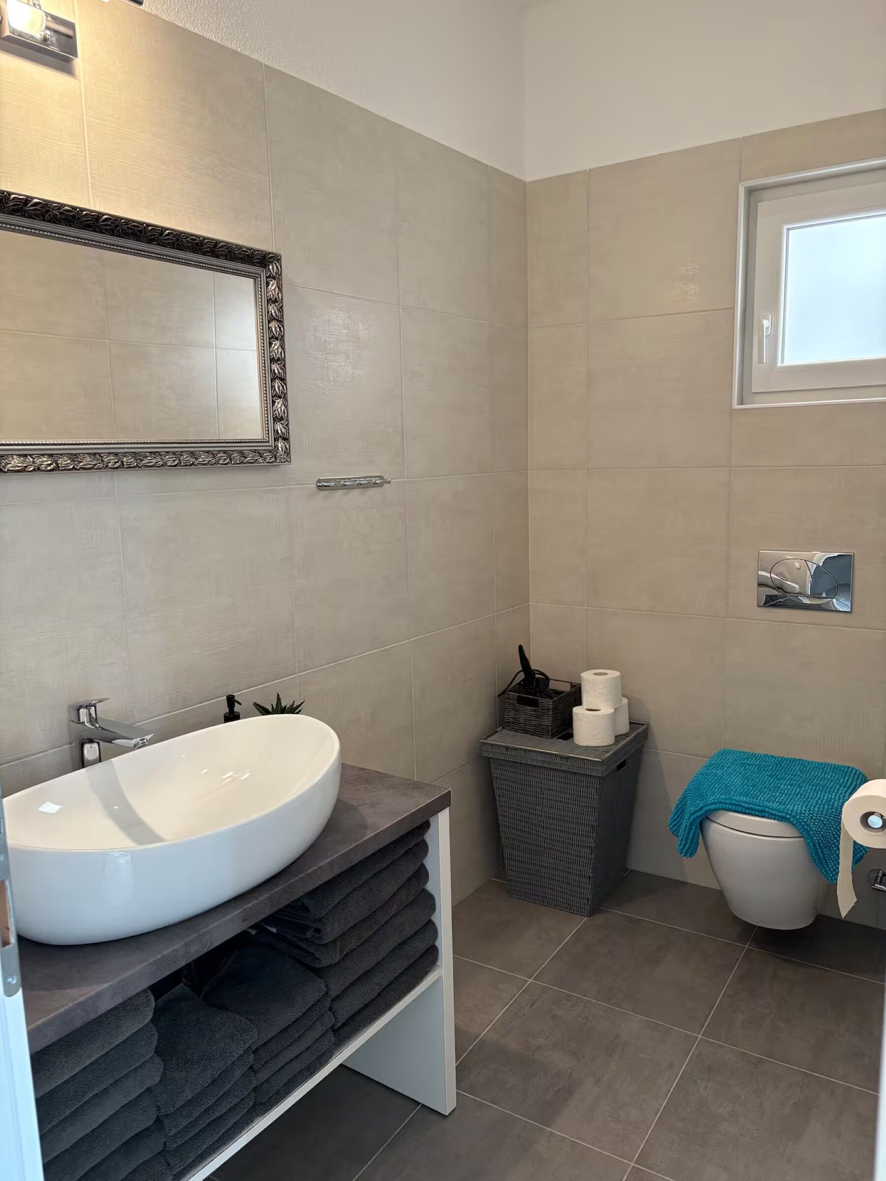 A bathroom with a large mirror, a white sink, a towel rack, a toilet, and a small window.