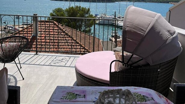 A terrace with wicker furniture, a table with a floral tablecloth, and a view of a lake with boats and hills in the background.