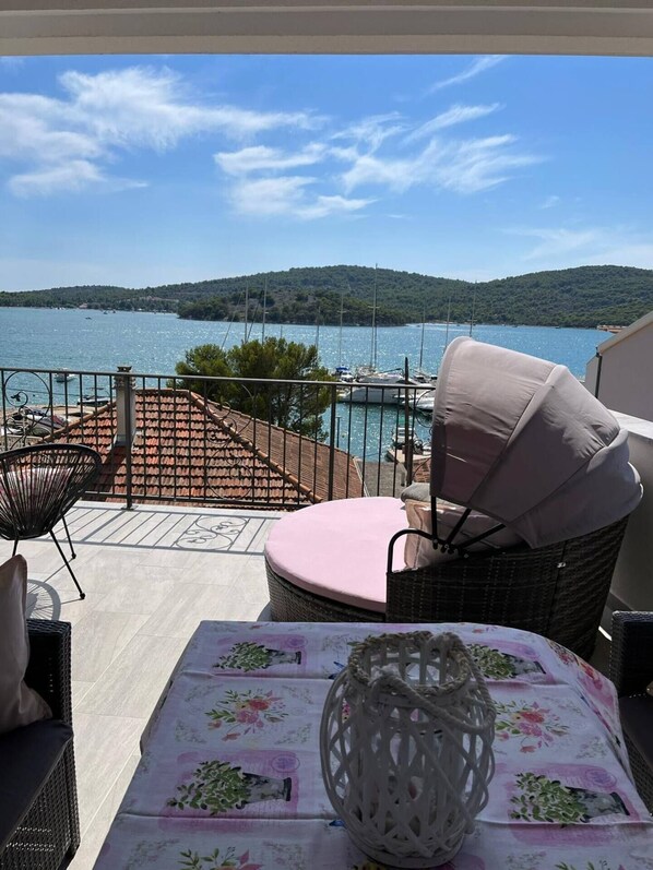 A terrace with wicker furniture, a table with a floral tablecloth, and a view of a lake with boats and hills in the background.