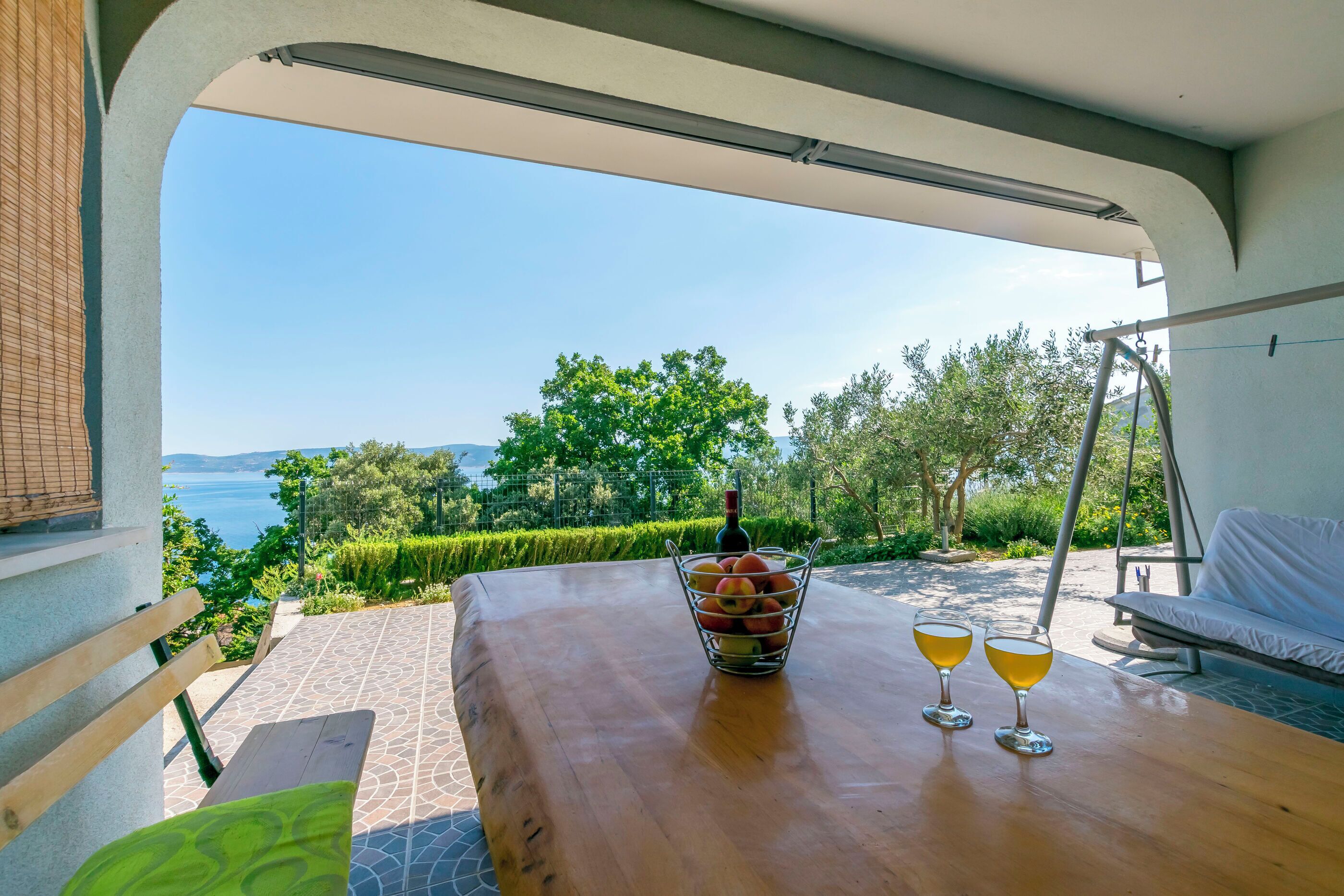 A wooden table with a fruit bowl and two glasses of wine, a white sofa, and a view of the sea and greenery.