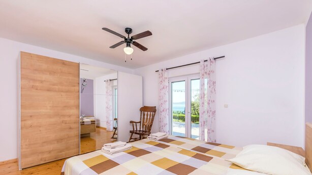 A bedroom with a bed, a ceiling fan, a chair, a window with curtains, and a wooden floor. - Apartment Perfect (Omis)