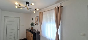 Ein Zimmer mit Tür, Schreibtisch mit Schubladen, Blumentopf, gerahmtes Bild, Leuchte und Fenster mit Vorhängen.