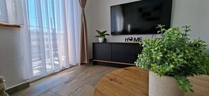 Ein Wohnzimmer mit an der Wand montiertem Fernseher, einem Topfpflanzen und Holzboden.