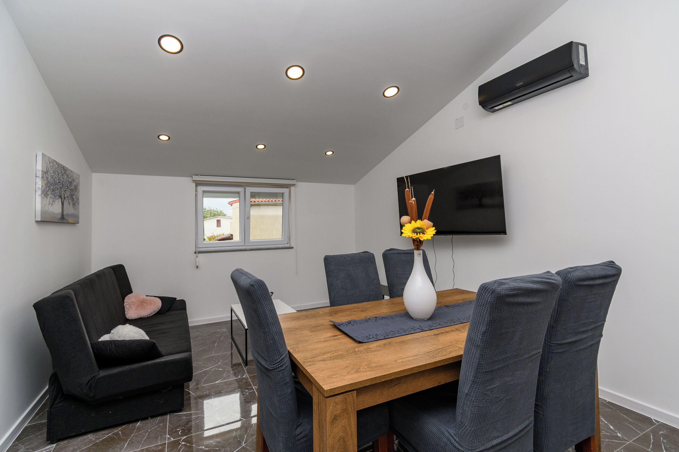 A modern dining area with a wooden table, grey upholstered chairs, a white vase with sunflowers, and a mounted flat-screen TV on the wall.