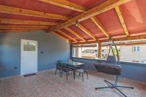 Eine überdachte Terrasse mit Tisch, Stühlen, einer Tür und Blick auf Gebäude durch das Fenster.
