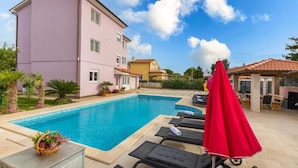 Ein Swimmingpool mit Liegestühlen und Sonnenschirmen, ein pinkfarbenes Gebäude und ein klarer Himmel.