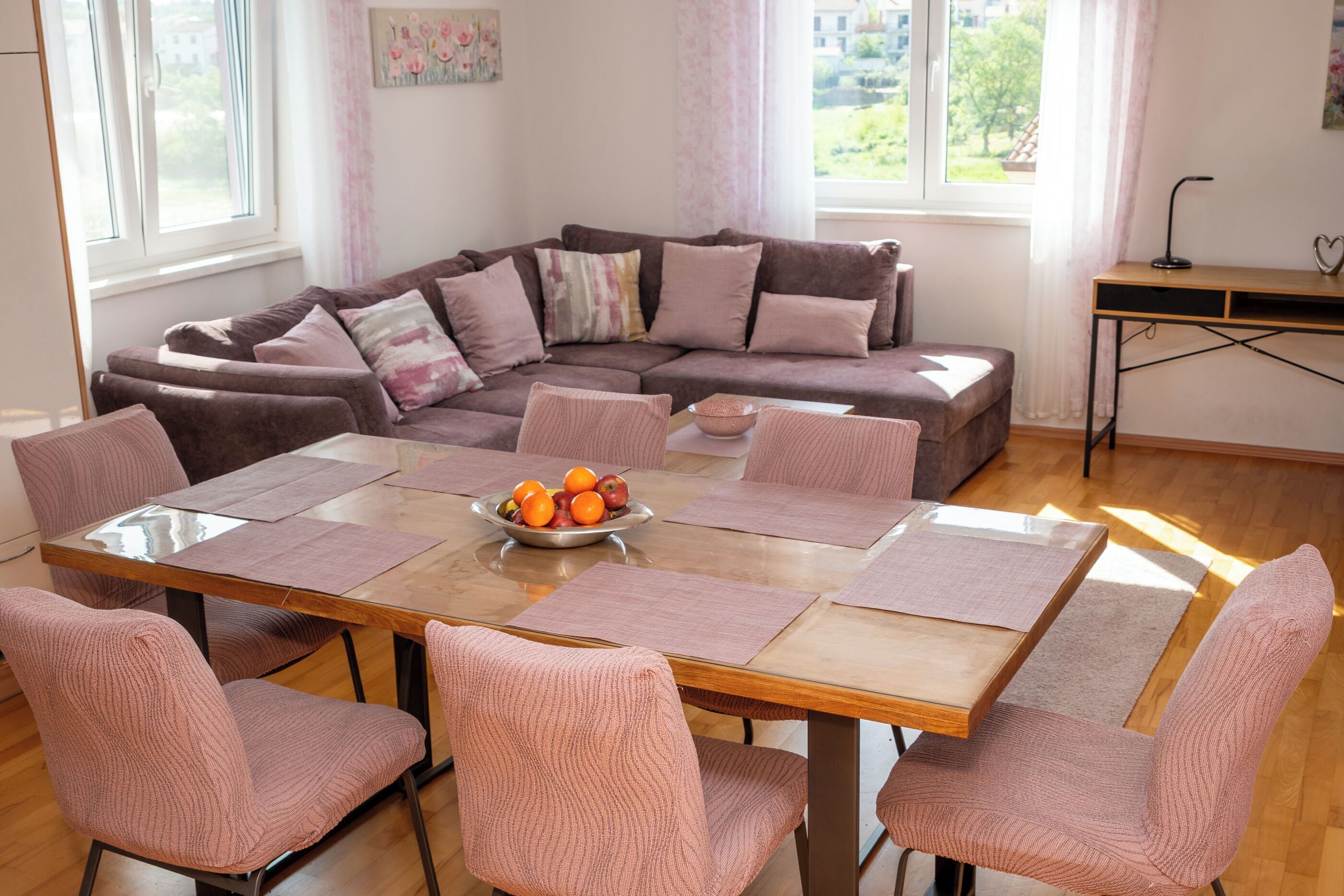 Ein gemütliches Wohnzimmer mit einer Eckcouch, einem Holzesstisch mit rosa Stühlen und einer Obstschale auf dem Tisch.