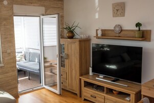 Un salón moderno con una puerta corrediza de vidrio que da a un balcón, un televisor de pantalla plana sobre un soporte de madera y una estantería con objetos decorativos.