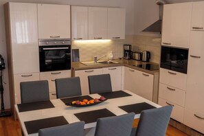 A modern kitchen with white cabinets, a built-in oven, a coffee maker, and a dining table with chairs. - Apartments Villa Ana (Fazana)